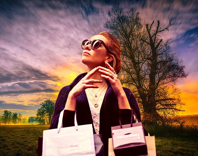 Eine stylische Frau mit Sonnenbrille steht im Freien, hält Einkaufstüten in beiden Händen und schaut nachdenklich in die Ferne. Im Hintergrund sind ein farbenfroher Sonnenuntergang und silhouettehafte Bäume zu sehen.