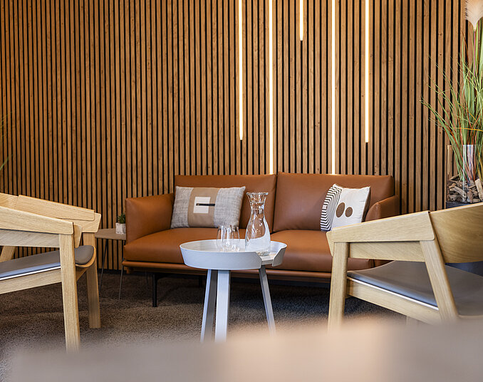 Moderne Sitzecke mit braunem Ledersofa, Holzsesseln, Wasserkaraffe und beleuchteter Lamellenwand