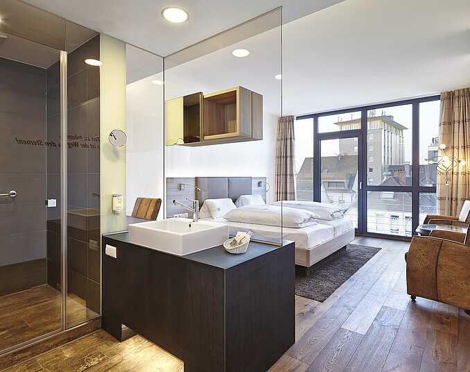 Modernes Hotelzimmer mit offenem Badezimmer, großem Doppelbett und bodentiefen Fenstern mit Stadtblick.