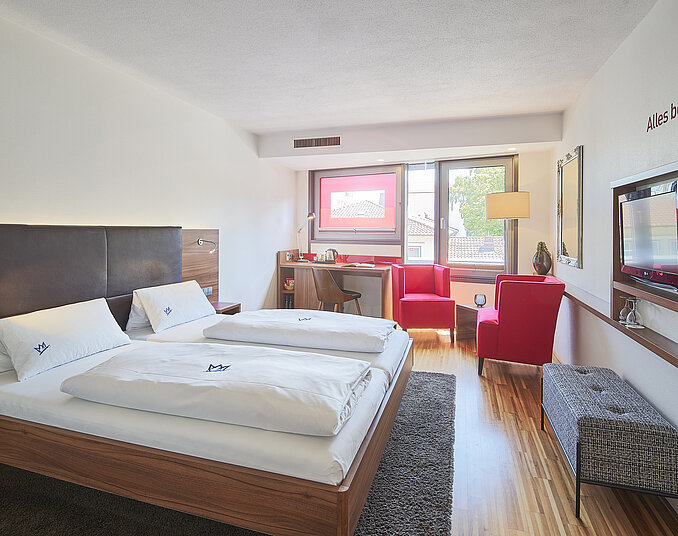 Das Bild zeigt ein modernes Hotelzimmer mit einem Doppelbett, einem Fenster mit Tageslicht, einem kleinen Tisch und zwei roten Sesseln. An der Wand ist ein Fernseher angebracht. Der Boden ist aus Holz und der Raum wirkt hell und einladend.