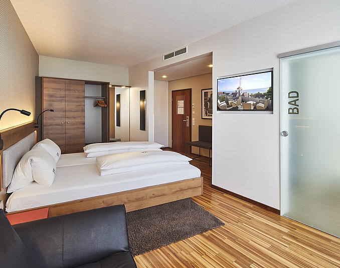 Ein modernes Hotelzimmer mit einem Doppelbett, einem schwarzen Sofa und einem Tisch. Die Wände sind in hellen Farben gehalten, und es gibt eine Glastür zur Badauswahl. Ein Holzregal und eine Lichtquelle über dem Bett sind ebenfalls sichtbar.