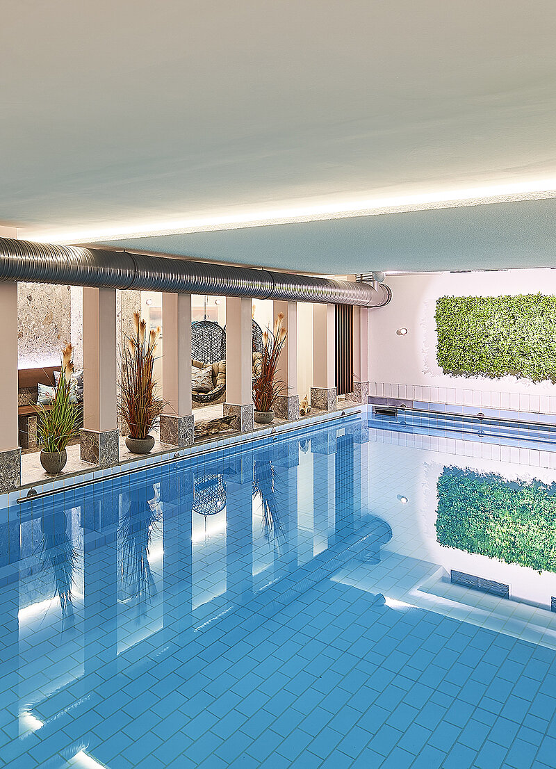 Ein moderner Innenpool mit glattem, blauem Wasser und einem hellen, einladenden Ambiente. An der Wand sind Pflanzen installiert, die zur entspannenden Atmosphäre beitragen. Die Beleuchtung sorgt für eine angenehme Stimmung im Raum.