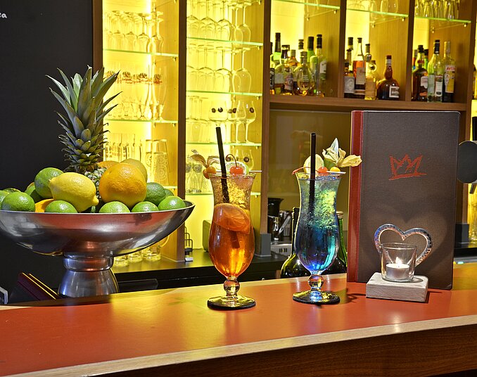 Die Bar zeigt eine Auswahl an bunten Cocktails in Gläsern, umgeben von frischen Früchten in einer Schale. Im Hintergrund sind Regale mit Spirituosen und Gläsern zu sehen. Ein Cocktail Menü ist ebenfalls sichtbar.