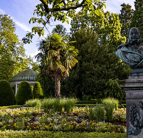 Sommerliche Gartenanlage mit Blumen und Palme in Friedrichshafen Prächtige Gartenanlage mit bunten Blumen, einer hohen Palme und einer historischen Büste, umgeben von üppigem Grün.