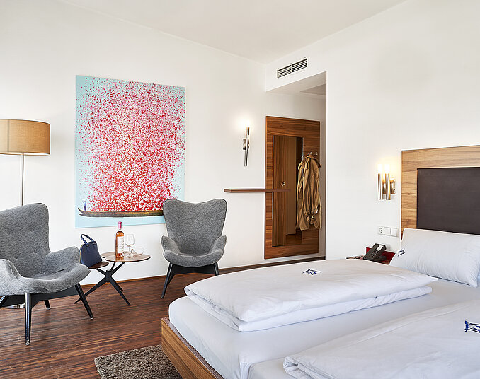 Ein modernes Hotelzimmer mit einem Doppelbett, zwei grauen Stühlen, einem kleinen Tisch und einem farbenfrohen Kunstwerk an der Wand. Die Einrichtung ist hell und minimalistisch, mit Holz- und Stoffelementen. Ein Kleiderschrank und eine Lampe sind ebenfalls sichtbar.
