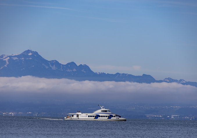Ein Passagierschiff fährt auf einem ruhigen See, während im Hintergrund die majestätischen Berge und eine Wolkenbank zu sehen sind. Der Himmel ist klar und hellblau.