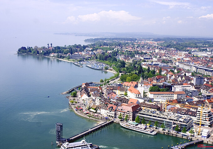 Luftaufnahme einer Küstenstadt am Bodensee, mit Aussicht auf Wohngebäude, Parks, Marina und Uferpromenade. Der Himmel ist blau mit einigen Wolken, und das Wasser reflektiert das Licht.