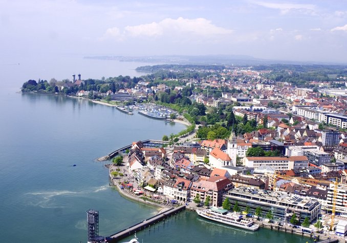 Luftaufnahme einer Küstenstadt am Bodensee, mit Aussicht auf Wohngebäude, Parks, Marina und Uferpromenade. Der Himmel ist blau mit einigen Wolken, und das Wasser reflektiert das Licht.