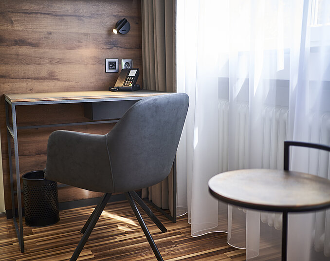 Ein moderner Arbeitsplatz mit einem grauen, komfortablen Stuhl an einem minimalistischen Schreibtisch. Im Hintergrund ist ein Telefon und eine Laminatoberfläche sichtbar. Neben dem Schreibtisch befindet sich ein kleiner runder Beistelltisch und ein Fenster mit Gardinen.