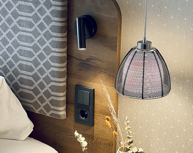 Detailaufnahme eines stilvollen Hotelbett-Kopfendes mit moderner Beleuchtung, Steckdose und Trockenblumen.