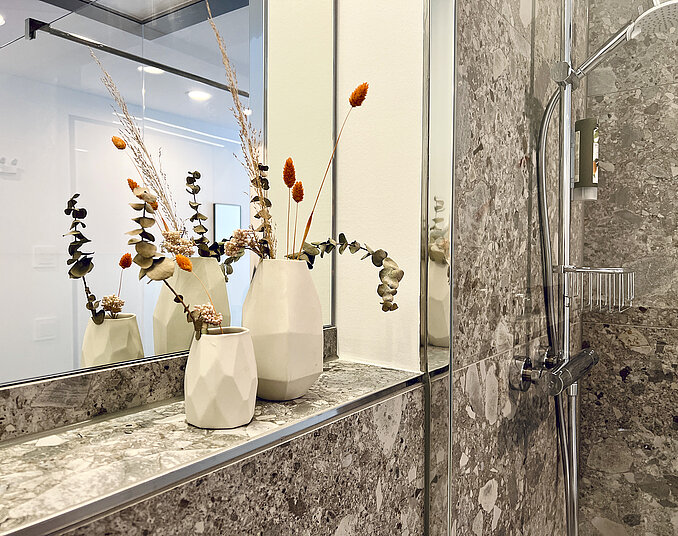 Stilvolles Badezimmer mit Natursteinfliesen, Spiegeln und weißen Vasen mit Trockenblumen als Dekoration.