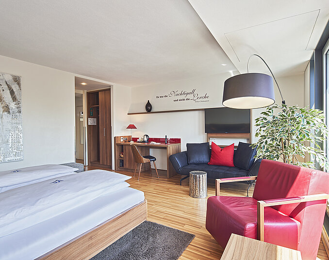 Ein modernes Hotelzimmer mit zwei Einzelbetten, einem bequemen Sitzbereich mit roten und blauen Kissen, einem Schreibtisch und einem großen Fenster. Eine Pflanze und eine stylische Stehleuchte ergänzen die gemütliche Atmosphäre. An der Wand ist eine dekorative Schrift angebracht.