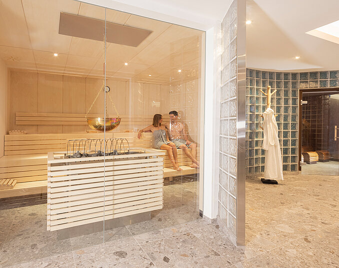 Paar in einer modernen Holzsauna mit Saunaofen, Aufgussschale und angrenzendem Wellnessbereich
