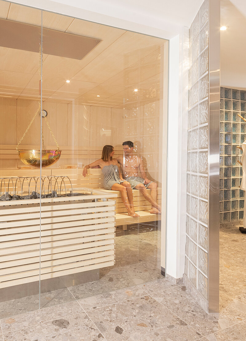 Paar in einer modernen Holzsauna mit Saunaofen, Aufgussschale und angrenzendem Wellnessbereich