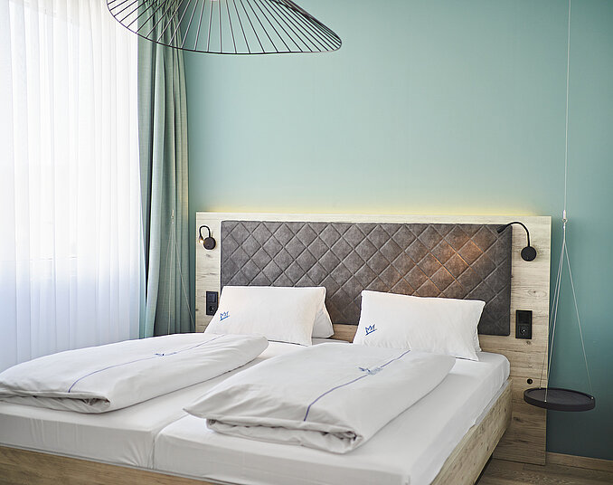 Schlafzimmer mit weißer Bettwäsche und einem gepolsterten Kopfteil. Eine große Fensterfront mit Vorhängen ist sichtbar, darüber hängt eine Drahtpendellampe. Die Wand ist in sanftem Grün gestrichen, und neben dem Bett sind Nachttischlampen angebracht.