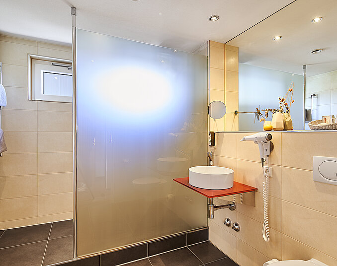 Helles modernes Badezimmer mit einer begehbaren Dusche, die durch eine matte Glaswand abgetrennt ist. Ein runder Waschbecken befindet sich auf einer roten Ablage, während an der Wand Handtücher hängen. Deckenleuchten sorgen für eine angenehme Beleuchtung.