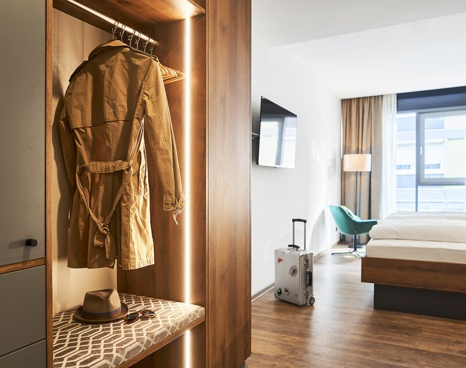 Modernes Hotelzimmer mit Doppelbett, offenem Kleiderschrank und bodentiefem Fenster mit Stadtblick.
