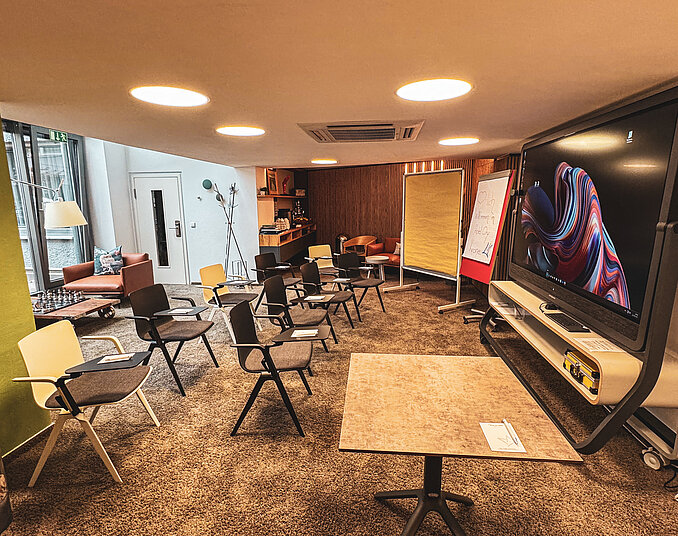 Ein moderner Raum mit einem großen Bildschirm, einem Tisch in der Mitte und mehreren Stühlen, die im Kreis angeordnet sind. Im Hintergrund sind eine Couch, ein Stehlampe und Flipcharts zu sehen. Der Boden ist mit Teppich ausgelegt und es gibt ausreichende Beleuchtung.