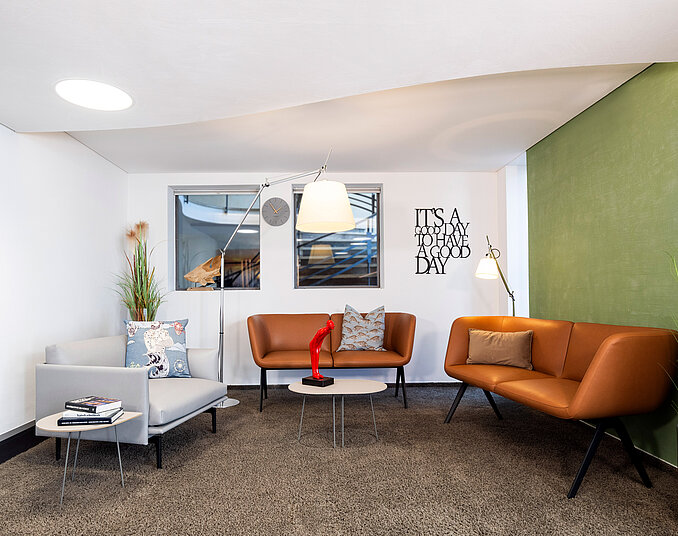 Gemütlicher Lounge-Bereich mit zwei modernen Sofas in Braun und Grau, einem runden Tisch und einer Stehleuchte. Eine grüne Wand und Pflanzen schaffen eine entspannte Atmosphäre. An der Wand hängt ein Spruch: "IT'S A GOOD DAY".