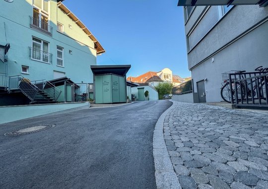 Blick auf eine schmale, asphaltierte Straße zwischen Gebäuden. Links stehen zwei Häuser mit Balkonen, rechts befindet sich ein moderner Gebäudeteil. Am Ende der Straße sind Fahrräder abgestellt und ein grüner Bereich ist sichtbar. Der Himmel ist klar und blau.