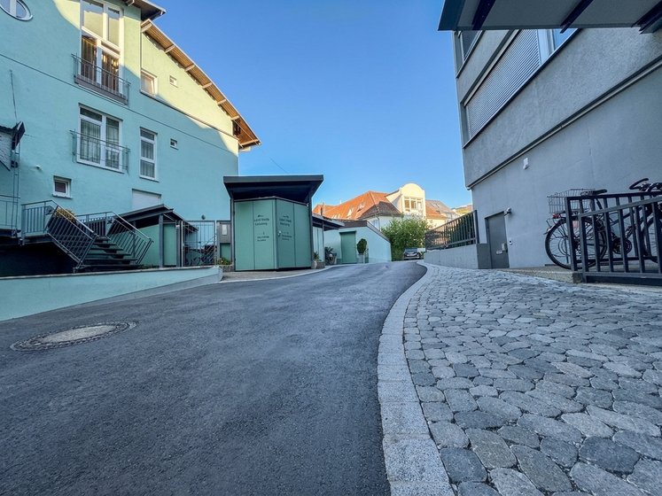 Blick auf eine schmale, asphaltierte Straße zwischen Gebäuden. Links stehen zwei Häuser mit Balkonen, rechts befindet sich ein moderner Gebäudeteil. Am Ende der Straße sind Fahrräder abgestellt und ein grüner Bereich ist sichtbar. Der Himmel ist klar und blau.