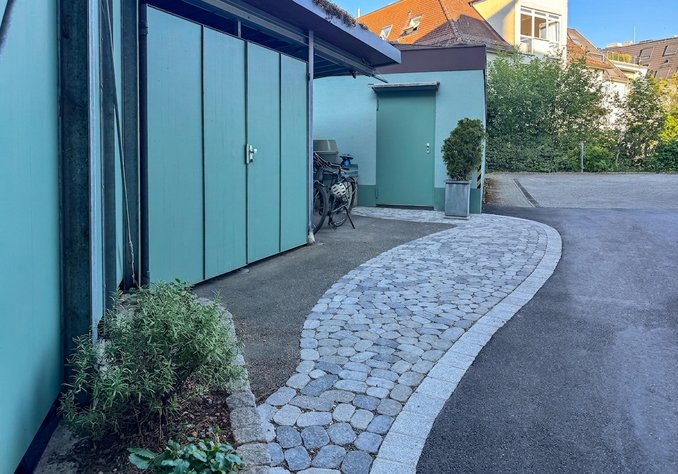 Ein gepflasterter Weg mit einer sanften Biegung führt zu einem kleinen Gebäude, das grüne Wände hat. Auf der linken Seite stehen einige Fahrradständer und eine niedrige Pflanze ist im Vordergrund sichtbar. Im Hintergrund sind Bäume und weitere Gebäude zu sehen.