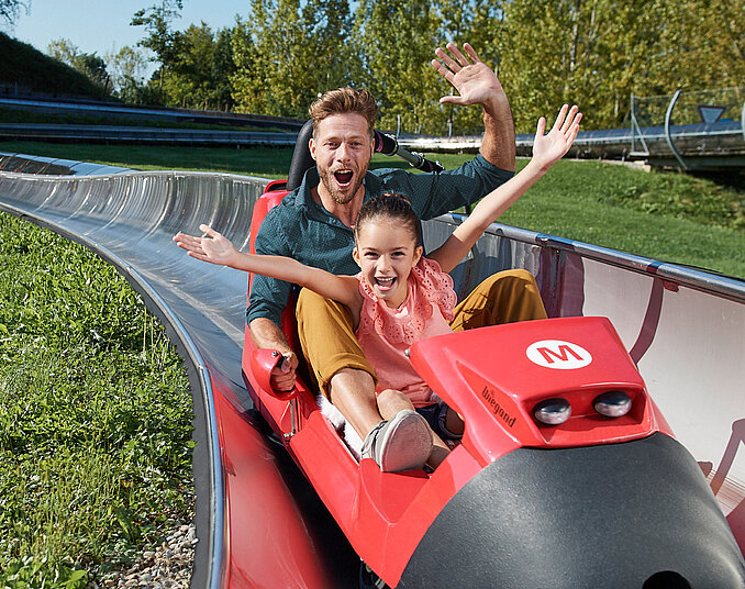 Ein Mann und ein Mädchen sitzen glücklich in einer roten Sommerrodelbahn. Beide haben die Arme in die Luft gehoben und lächeln, während sie rasch den Hang hinunterfahren. Im Hintergrund sind grüne Bäume und eine Rodelbahn zu sehen.