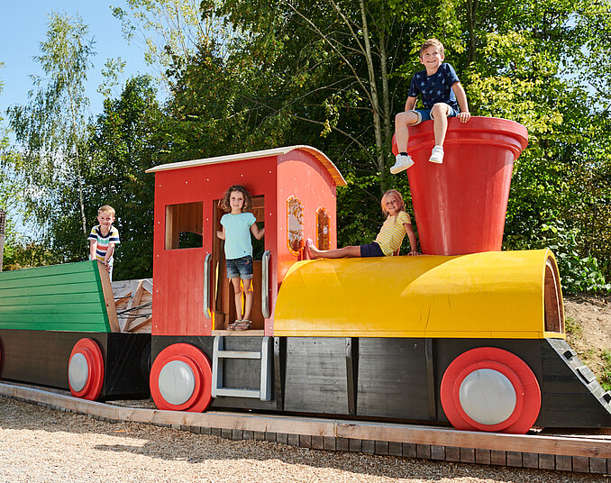 Drei Kinder spielen auf einem bunten Spielzeugzug im Freien. Zwei Kinder sitzen auf dem Zug, während ein weiteres Kind neben dem Zug steht. Der Zug hat einen roten Führerstand und eine gelbe Lokomotive mit großen Rädern. Im Hintergrund sind Bäume und grünes Gemüse sichtbar.