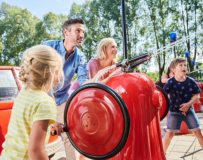 Eine Familie mit zwei Erwachsenen und zwei Kindern spielt an einem Wasserspielplatz. Sie bedienen rote Wasserpumpen, während Wasser spritzt. Im Hintergrund sind Bäume und Spielgeräte zu sehen. Sonne scheint und die Stimmung ist fröhlich.