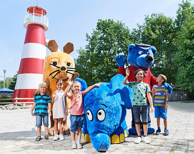 Sechs Kinder stehen fröhlich vor einem roten und weißen Leuchtturm. Neben ihnen stehen große, bunte Kostüme von einem Bären, einer Maus und einem blauen Elefanten. Alle lächeln und haben Spaß in einer sonnigen Umgebung.