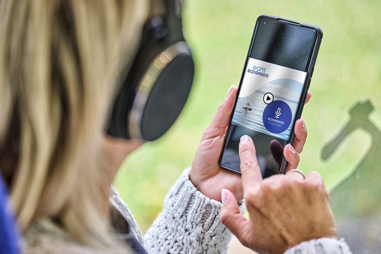 Eine Person mit Kopfhörern hält ein Smartphone in der Hand, auf dem eine App angezeigt wird. Die App zeigt eine Vorschau für eine Audioaufnahme mit einem Mikrofon-Symbol und einer Schaltfläche zum Abspielen an. Der Hintergrund ist unscharf und zeigt eine natürliche Umgebung.