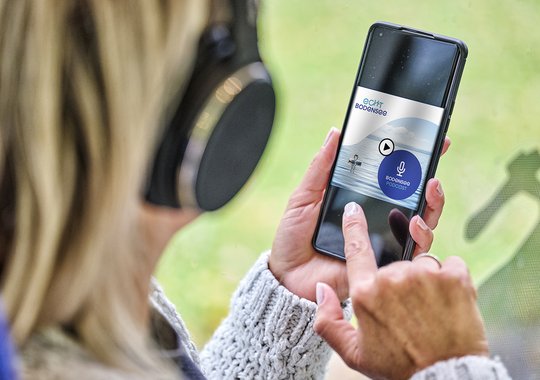 Eine Person mit Kopfhörern hält ein Smartphone in der Hand, auf dem eine App angezeigt wird. Die App zeigt eine Vorschau für eine Audioaufnahme mit einem Mikrofon-Symbol und einer Schaltfläche zum Abspielen an. Der Hintergrund ist unscharf und zeigt eine natürliche Umgebung.