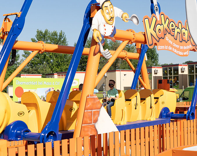 Ein bunter Freizeitpark-Attraktion namens "Kakerlaken" ist zu sehen. Sie besteht aus einer gelben, insektähnlichen Bahn mit einem fröhlichen Koch in einem weißen Outfit, der darauf steht. Die Bahn ist von einer orangefarbenen Umzäunung umgeben und in einem sonnigen Außenbereich platziert.