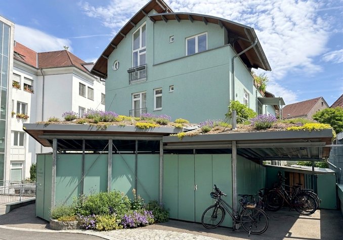 Ein modernes Wohnhaus mit grün gestrichener Fassade und einem begrünten Dach. Darunter befindet sich ein überdachter Fahrradabstellplatz mit mehreren Fahrrädern. Die Umgebung ist hell und freundlich, mit blauem Himmel und ein paar Wolken.